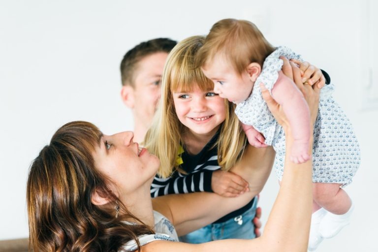 Une femme porte joyeusement un bébé dans une robe claire tandis qu'une jeune fille aux cheveux blonds, vêtue d'une chemise rayée, sourit au bébé. On aperçoit partiellement un homme derrière eux, souriant chaleureusement. Le fond blanc uni souligne le bonheur de la famille et met en valeur ces moments simples en famille.