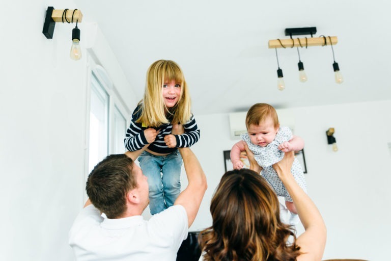 Un homme et une femme, tous deux tournés vers l'objectif, portent joyeusement deux enfants dans une pièce lumineuse. L'homme tient une fillette souriante aux cheveux blonds et portant une chemise rayée, tandis que la femme soulève un bébé vêtu d'une tenue blanche. La pièce est dotée de luminaires modernes et de murs blancs, capturant des moments simples en famille.