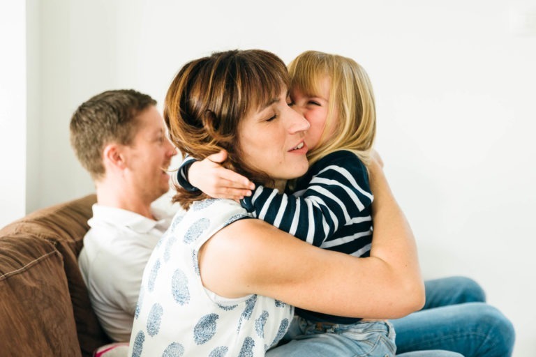 Une femme aux cheveux bruns courts serre dans ses bras une jeune fille souriante aux cheveux blonds et portant une chemise rayée, toutes deux assises sur un canapé. En arrière-plan, un homme aux cheveux courts et portant une chemise blanche est assis, les regardant avec un sourire. Le décor semble être un environnement familial détendu, capturant des moments simples en famille.