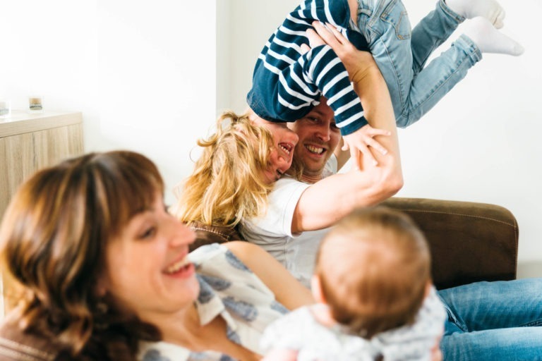 Un moment de joie en famille avec quatre membres dans un salon chaleureux et bien éclairé. Un père soulève joyeusement un bambin rieur la tête en bas, tandis qu'une mère, souriante, tient un bébé dans ses bras. La famille semble détendue et heureuse, profitant de ces moments simples en famille.