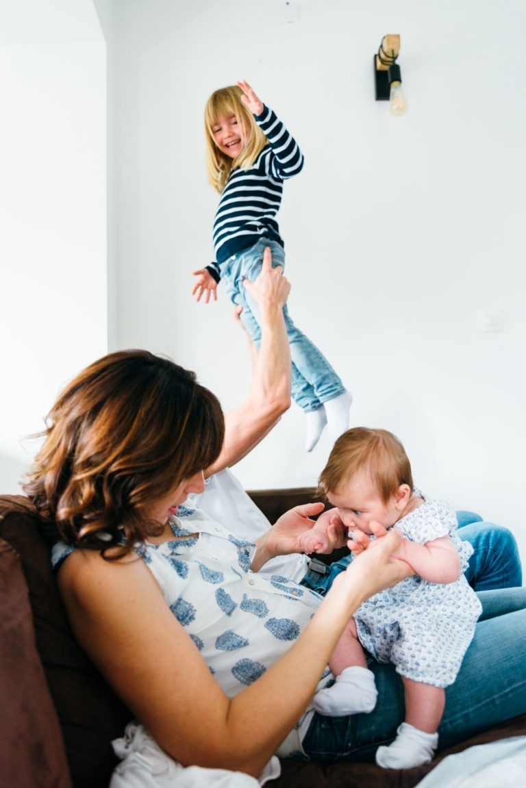 Une scène familiale capture des moments simples en famille, avec une mère tenant un bébé dans une robe blanche et bleue sur ses genoux. La mère, assise sur un canapé marron, regarde avec amour quelqu'un soulever dans les airs un jeune enfant portant une chemise rayée et un jean. L'enfant est excité, le bras levé et sourit largement.