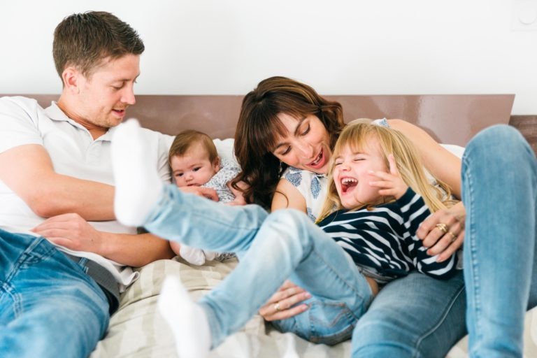 Une famille heureuse est assise sur un lit. Le père, vêtu d'une chemise blanche, et la mère, aux longs cheveux bruns, tiennent un bébé dans leurs bras. Un enfant plus âgé, vêtu d'une chemise rayée et d'un jean, rigole tandis que la mère lui chatouille les côtes. Ces moments simples en famille sont remplis de joie et de rires.