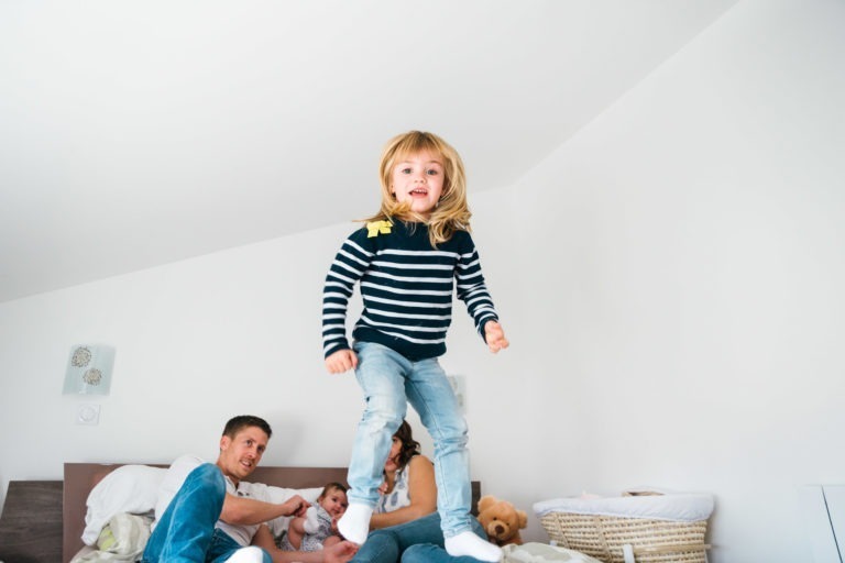 Un jeune enfant aux longs cheveux blonds, vêtu d'une chemise rayée et d'un jean bleu, saute joyeusement sur un lit. Un couple et un autre jeune enfant sont assis à l'arrière-plan sur le lit, souriant et riant. La scène capture des moments simples en famille dans une pièce lumineuse et aérée aux murs blancs et au décor minimaliste.