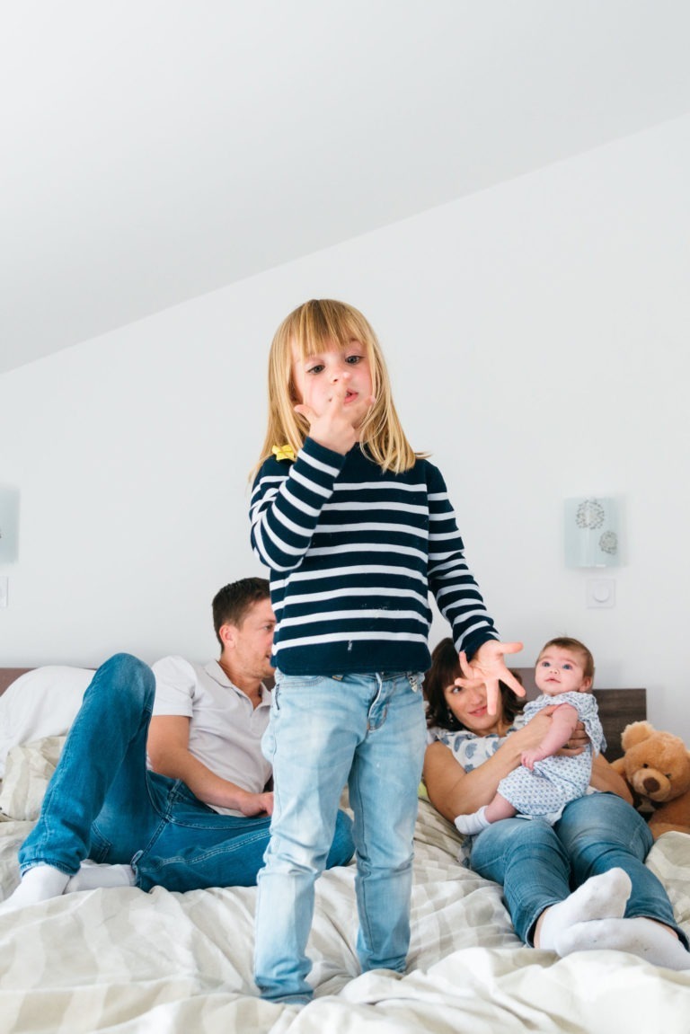 Une jeune fille blonde portant un pull rayé noir et blanc et un jean bleu se tient debout sur un lit, se couvrant la bouche avec sa main. Derrière elle, un homme et une femme sont allongés sur le lit, la femme tenant un bébé habillé en blanc. Un ours en peluche est placé à côté de la famille, capturant des moments simples en famille.