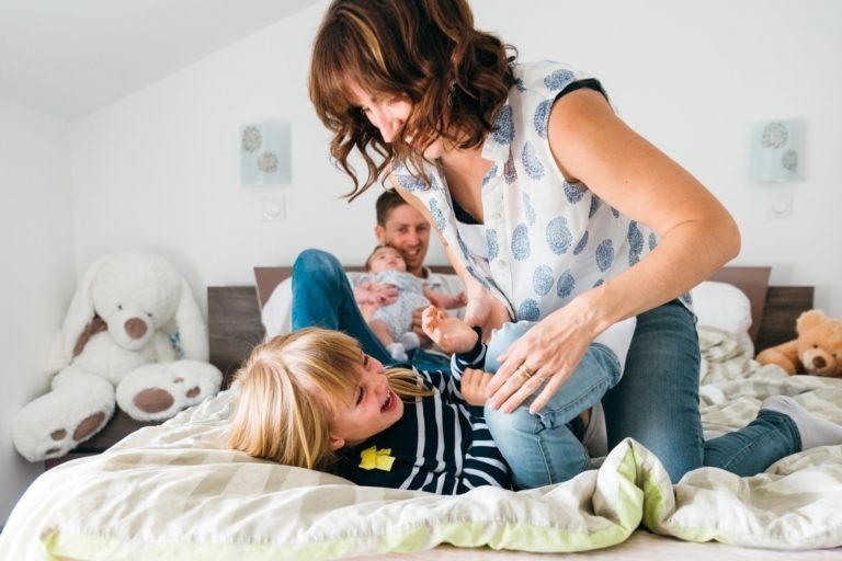 Une femme joue joyeusement avec une jeune fille sur un lit tandis qu'un homme est allongé à l'arrière-plan, tenant un bébé dans ses bras. La pièce est décorée de gros animaux en peluche. La scène dépeint des moments simples en famille, remplis de rires et d'interactions ludiques.