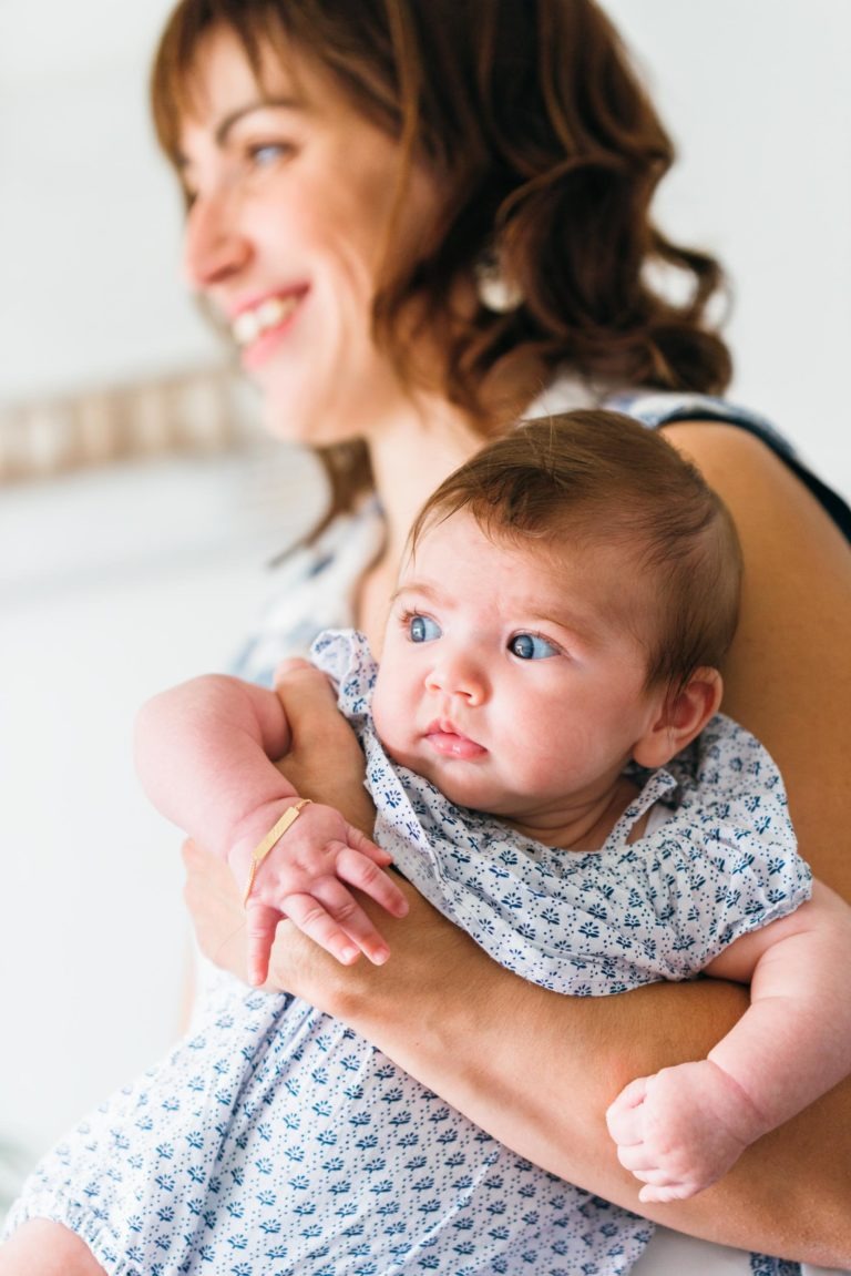 Une femme aux cheveux châtains mi-longs, souriante et regardant au loin, tient dans ses bras un bébé alerte aux yeux bleus. Le bébé est vêtu d'une combinaison blanche à motifs bleus et porte un bracelet en or au poignet gauche. L'arrière-plan doucement flouté met en valeur ces moments simples en famille, en mettant l'accent sur la femme et le bébé.