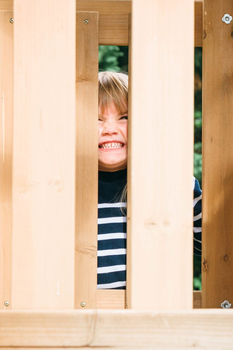 Un enfant aux cheveux blonds et à la chemise rayée sourit largement, jetant un œil à travers les interstices verticaux d'une clôture en bois. La clôture marron clair, avec des vis visibles aux coins, encadre cet instantané parfait de moments simples en famille. La verdure floue en arrière-plan évoque un cadre extérieur idyllique.