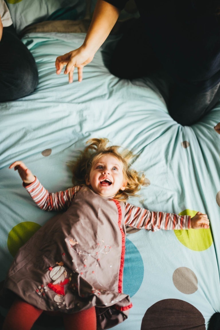 Une petite fille aux cheveux blonds est allongée sur un lit, souriant joyeusement, les bras tendus au-dessus de sa tête. Elle porte une chemise rayée et une robe de Maison du Bonheur avec un motif de vacances. La main d'un adulte se tend vers l'enfant depuis le haut de l'image. Le couvre-lit a un motif à pois.