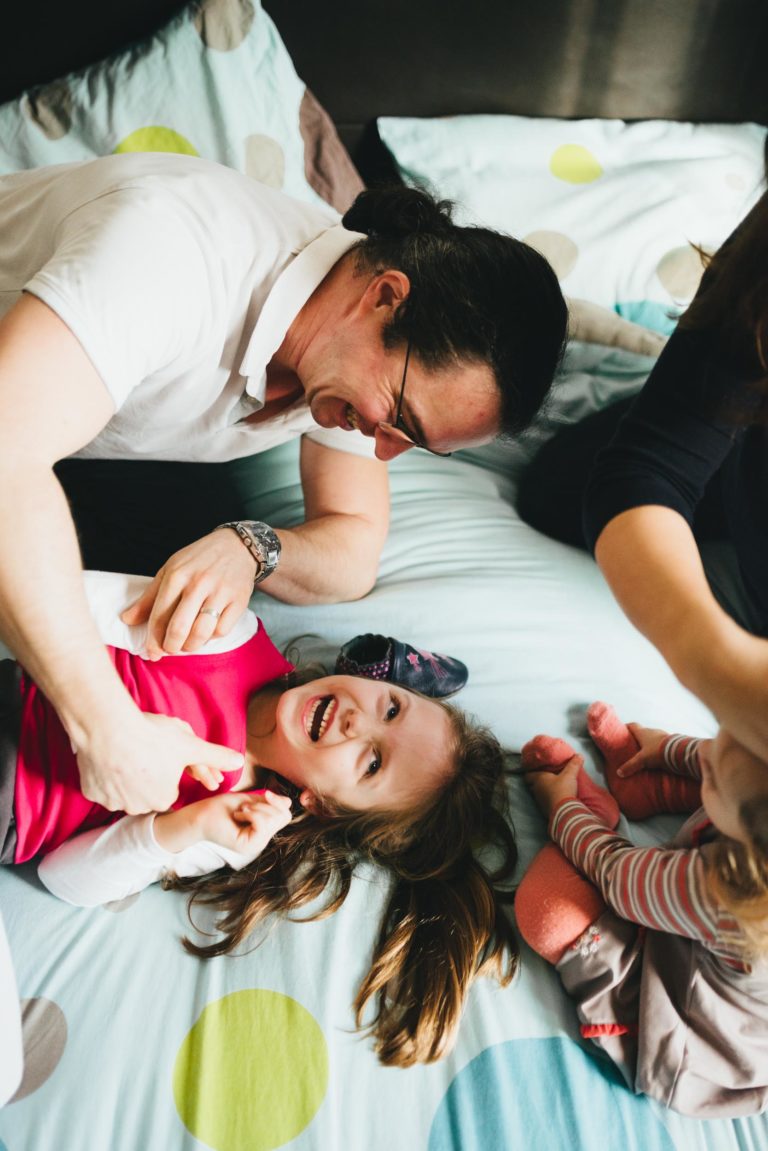 Un homme à lunettes chatouille de manière ludique une jeune fille allongée sur un lit de la Maison du Bonheur. La fille, qui sourit largement dans sa chemise rose, est à côté d'un autre enfant en vêtements rayés et chaussettes roses. Le lit à pois colorés ajoute à l'ambiance joyeuse, ainsi que la présence d'un autre adulte à proximité.