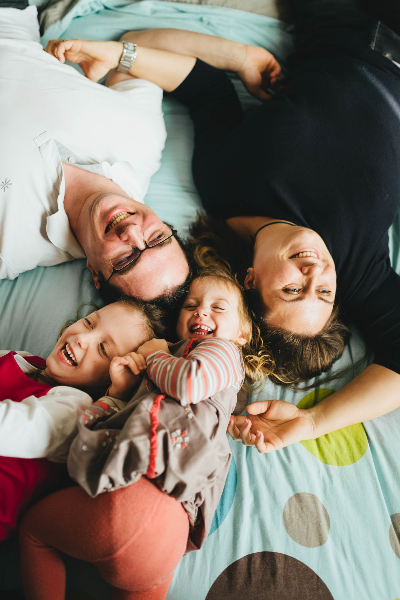 Une joyeuse famille de quatre personnes est allongée sur un lit. Deux parents sourient allongés sur le dos, avec deux jeunes enfants blottis entre eux, tous deux riant. Le couvre-lit Maison du Bonheur est bleu clair avec un motif de cercles de différentes tailles en vert, marron et blanc.