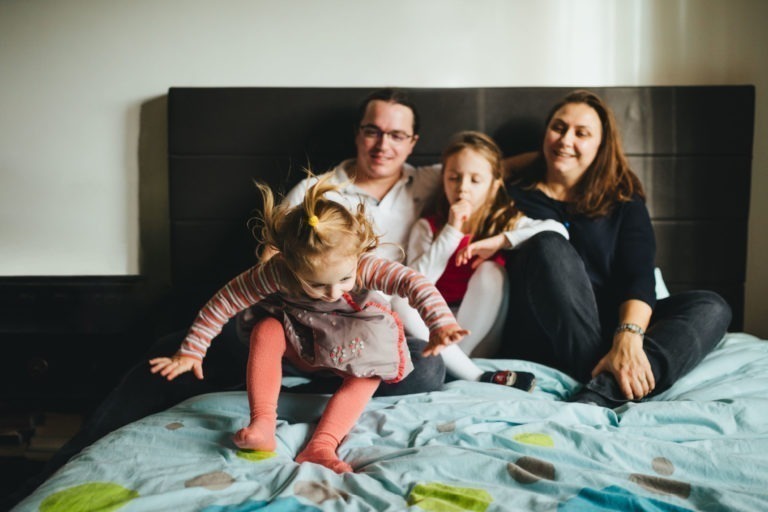 Une jeune famille est assise sur un lit à pois bleus à la Maison du Bonheur. Un homme et une femme adultes sourient, assis derrière deux enfants. L'un d'eux est une fille aux nattes blondes portant une chemise rayée et des leggings roses, sautant sur le lit, tandis que l'autre fille aux cheveux longs regarde avec un doigt près de sa bouche.