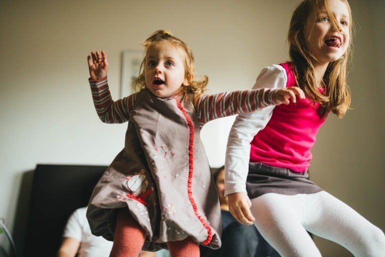 Deux jeunes filles sautent joyeusement à l'intérieur de la Maison du Bonheur. La plus jeune à gauche porte une robe grise, tandis que la plus âgée à droite porte un haut rouge et des collants blancs. Toutes deux affichent des expressions d'excitation et de bonheur, l'arrière-plan de la pièce étant légèrement flou.