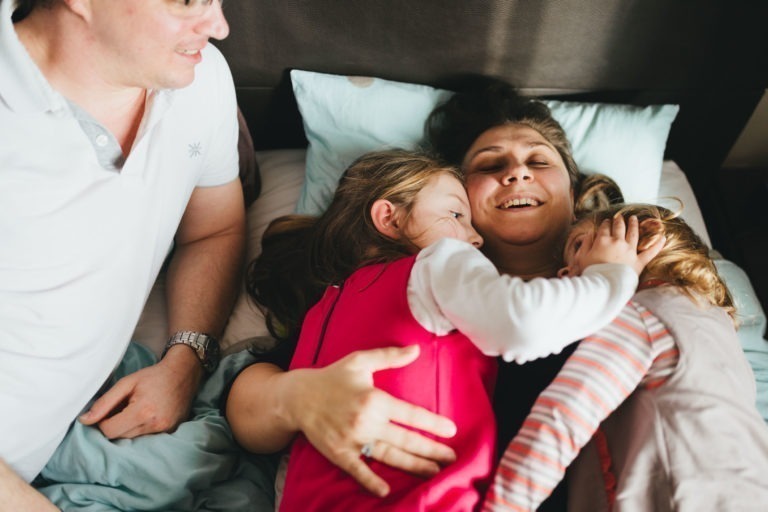 Une femme allongée sur un lit serre joyeusement dans ses bras deux enfants qui sont également allongés sur elle. L'un des enfants a de longs cheveux bruns et porte un haut rose et des manches blanches, tandis que l'autre enfant a les cheveux plus courts et des manches rayées. Un homme, portant des lunettes et un polo blanc, est assis à proximité et sourit dans leur Maison du Bonheur.