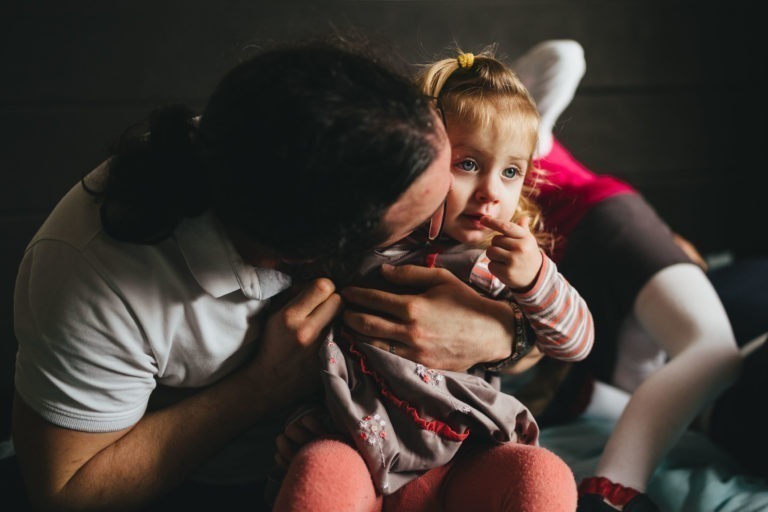 Une personne portant des lunettes tient et embrasse affectueusement la joue d'un petit enfant qui suce son pouce. L'enfant, vêtue d'une chemise rayée sous une robe à motifs floraux, a les cheveux blonds attachés par un ruban jaune. Dans ce moment de tendresse à la Maison du Bonheur, d'autres personnes sont partiellement visibles à l'arrière-plan.