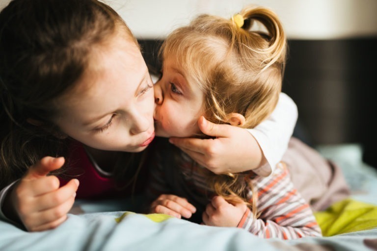 Deux jeunes filles partagent un moment d'affection à l'intérieur. La fille aînée est allongée sur le côté tandis que la plus jeune, aux cheveux blonds attachés en queue de cheval, l'embrasse sur la joue. Elles sourient toutes les deux tandis que la fille aînée tient doucement le visage de la plus jeune. La lumière naturelle de la Maison du Bonheur illumine la scène.