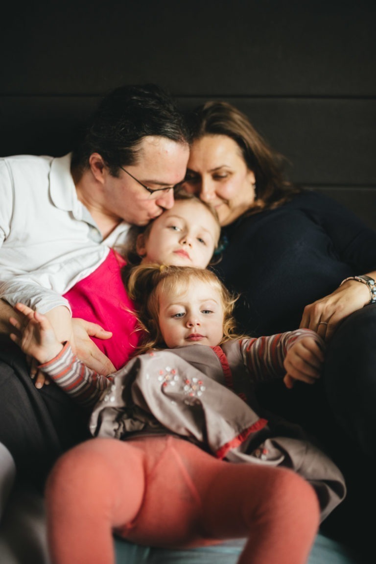 Une famille de quatre personnes est allongée côte à côte sur un canapé dans leur confortable Maison du Bonheur. L'homme et la femme aux cheveux noirs enlacent leurs deux enfants : celui en chemise rose est blotti entre les adultes, tandis que le plus jeune, vêtu d'une robe à motifs et d'un legging rose, se détend tranquillement devant.