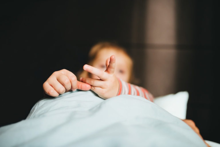 Gros plan d'un enfant allongé dans son lit, recouvert en grande partie d'une couverture bleu clair de Maison du Bonheur. L'accent est mis sur les mains de l'enfant qui sortent de sous la couverture, une main jouant avec les doigts de l'autre. L'enfant porte une chemise à manches longues rayée. L'arrière-plan est sombre et flou.