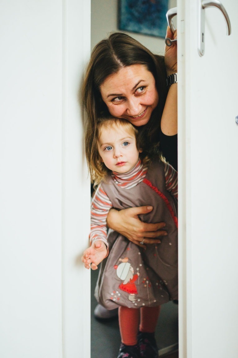 Une femme au sourire chaleureux et une jeune fille à l'expression sérieuse regardent par une porte blanche entrouverte de la Maison du Bonheur. La jeune fille porte une robe grise à motifs rouges et blancs et une chemise rayée à manches longues. La femme tient la porte et se penche, son visage près de la tête de la jeune fille.