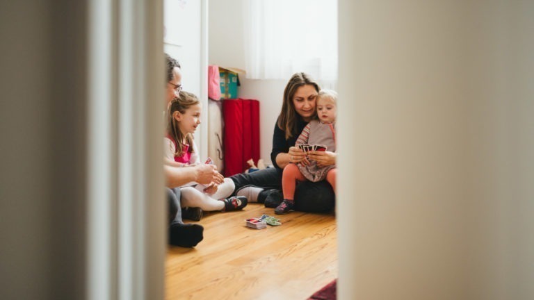 Une vue à travers une porte de la Maison du Bonheur montre une famille de quatre personnes assises sur un plancher en bois dans une pièce lumineuse. La mère, tenant un bébé, est entourée du père et d'un autre enfant, jouant avec un jeu de cartes. Des jouets et des bacs de rangement colorés sont visibles à l'arrière-plan.