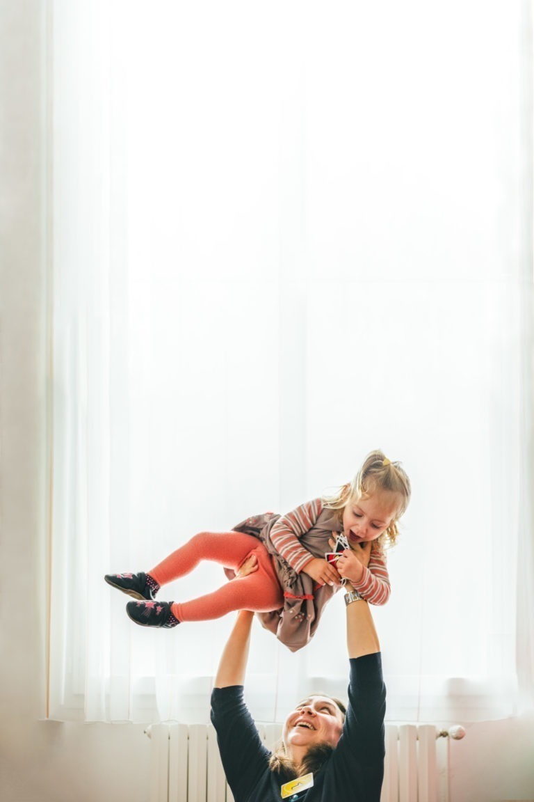 Un adulte, partiellement visible en bas, soulève dans les airs une jeune fille souriante portant une chemise rayée, des collants orange et des chaussures noires. La fille regarde vers le bas et semble ravie. Ils se trouvent dans une pièce très éclairée de la Maison du Bonheur avec une grande fenêtre recouverte de rideaux blancs transparents.