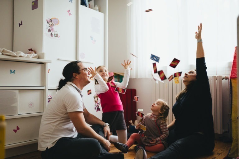 Une famille de quatre personnes est assise par terre dans une chambre et lance joyeusement des cartes à jouer en l'air. La pièce de leur Maison du Bonheur est dotée de meubles blancs, d'autocollants muraux colorés et d'une fenêtre lumineuse avec des rideaux transparents. Les parents et les deux enfants sourient et rient, créant une atmosphère animée et joyeuse.