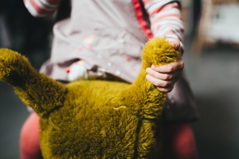 Un enfant vêtu d'une tenue rose et grise, aux manches rayées, tient l'oreille verte et duveteuse d'une peluche de la Maison du Bonheur. La peluche est partiellement visible au premier plan et semble douce et grande. L'image se concentre sur la main de l'enfant qui saisit l'oreille du jouet.