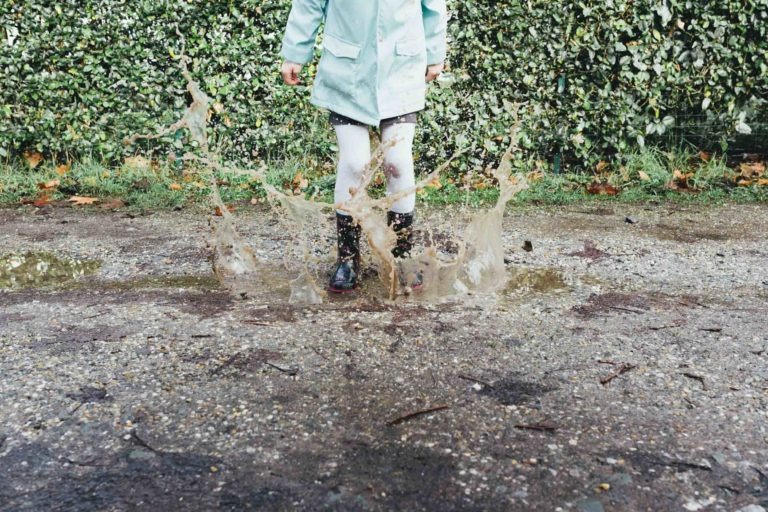 Une personne portant un manteau bleu clair, un pantalon blanc et des bottes noires est représentée en plein saut dans une flaque boueuse. Des éclaboussures d'eau sont capturées autour de ses jambes. Le décor de la scène ressemble aux charmants jardins de la Maison du Bonheur, avec une haie verte et des feuilles d'automne éparpillées sur le sol.