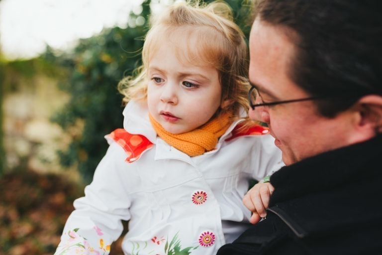 Un jeune enfant aux cheveux clairs et à la veste blanche aux motifs colorés est tenu par un adulte portant des lunettes et une veste noire. L'enfant, l'air pensif, regarde légèrement vers la gauche. L'arrière-plan, flou mais débordant de verdure, évoque l'atmosphère sereine de la Maison du Bonheur.