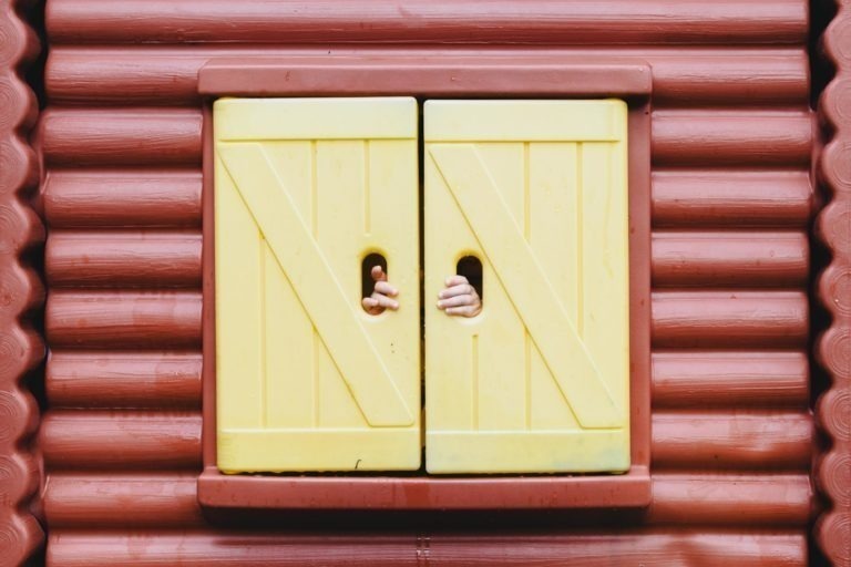 Deux petites mains regardent à travers les trous des volets jaunes fermés, posés sur un mur rouge de style cabane en rondins. Les volets de la Maison du Bonheur ont un design simple avec des lignes diagonales, suggérant que quelqu'un se trouve à l'intérieur de la structure.