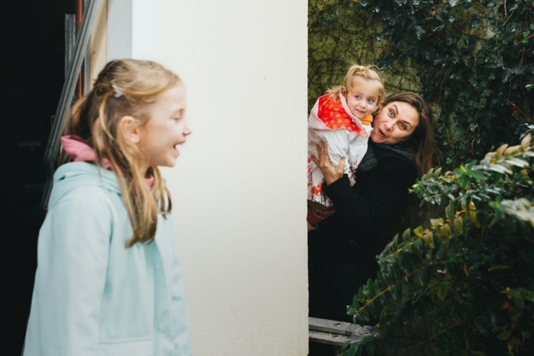 Au premier plan, une jeune fille en manteau bleu clair et capuche rose se tient en train de rire. Derrière un mur blanc de la Maison du Bonheur, une femme aux cheveux bruns se penche, tenant dans ses bras un bambin souriant dans une veste à capuche rouge ornée de motifs blancs. Un feuillage vert entoure la scène, ajoutant une touche de nature.