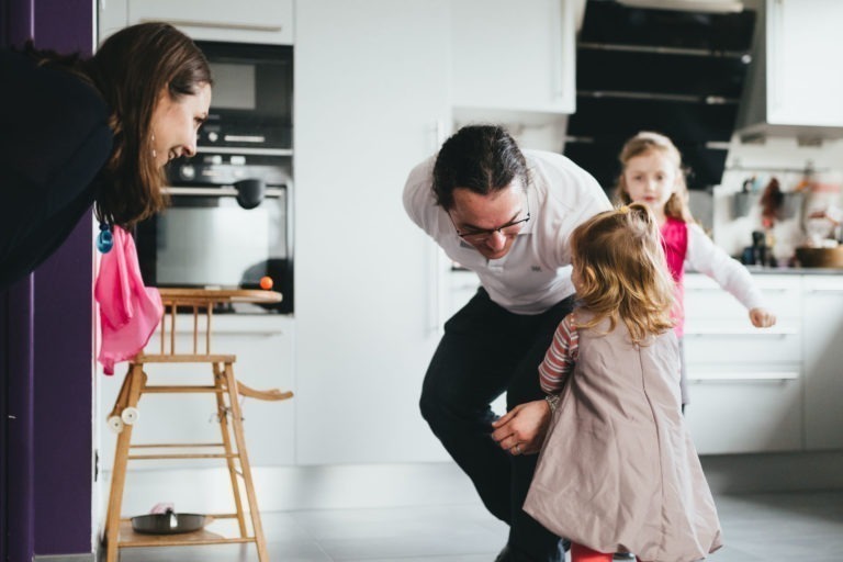 Une femme souriante se tient debout à côté d'une chaise haute en bois dans une cuisine moderne, observant un homme agenouillé et souriant à une jeune fille en robe grise. Un autre enfant, portant un foulard rose, se tient à proximité. La scène est lumineuse et familiale, incarnant le charme de la Maison du Bonheur avec des armoires de cuisine et des appareils électroménagers blancs en arrière-plan.