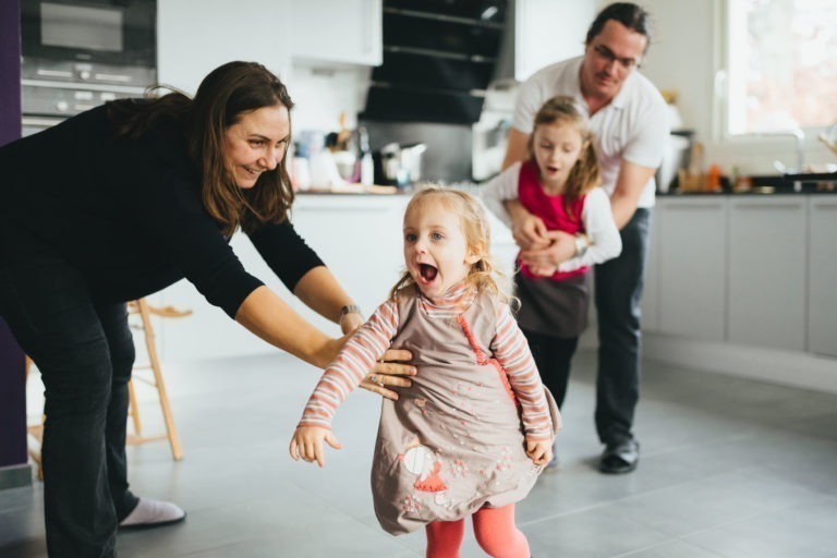 Un moment de joie en famille se déroule dans la cuisine de la Maison du Bonheur. Deux adultes jouent avec deux enfants, l'un court en riant et l'autre est doucement guidé par un adulte. Le cadre est décontracté avec une cuisine moderne en arrière-plan et tout le monde sourit.