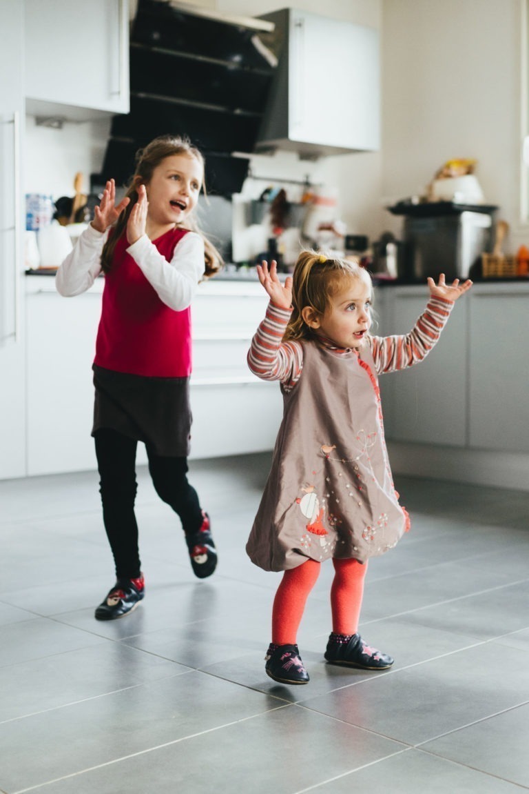 Deux jeunes filles dansent joyeusement dans une cuisine moderne, rappelant une Maison du Bonheur. La fille au premier plan porte une robe grise avec des collants roses et des manches rayées, elle lève les mains joyeusement. Derrière elle, une autre fille en haut rouge et jupe noire applaudit avec enthousiasme. La cuisine est lumineuse avec des meubles de couleur claire.