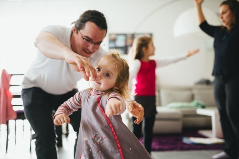 Un adulte portant des lunettes aide un jeune enfant vêtu d'une robe grise et d'une chemise rayée à se tenir en équilibre tout en tendant les bras. Deux autres enfants à l'arrière-plan tendent également les bras. La scène, remplie de lumière naturelle et de meubles, capture un moment de joie dans la chaleureuse Maison du Bonheur.