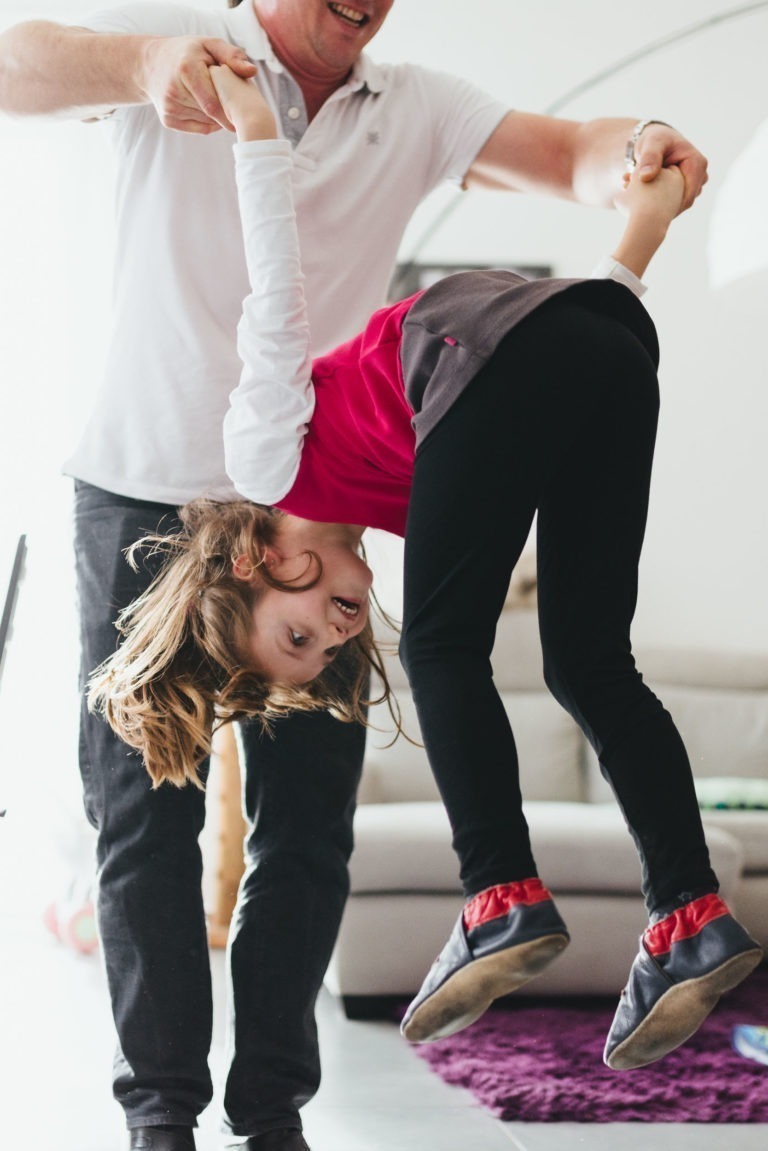 Un enfant vêtu d'un pull rouge et blanc et d'un pantalon noir est joué par un adulte vêtu d'une chemise blanche et d'un pantalon foncé, qui lui tient les mains. L'enfant sourit et regarde la caméra. Ils sont à l'intérieur de la Maison du Bonheur, avec un canapé beige et un tapis violet visibles en arrière-plan.