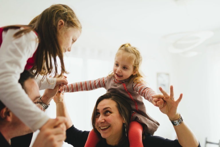 Une femme adulte souriante porte une jeune fille sur ses épaules à l'intérieur. La jeune fille, souriante elle aussi, tient la main d'un autre enfant qui se penche vers elle. La scène à la Maison du Bonheur est lumineuse et joyeuse, avec la lumière du soleil qui entre par les fenêtres en arrière-plan.