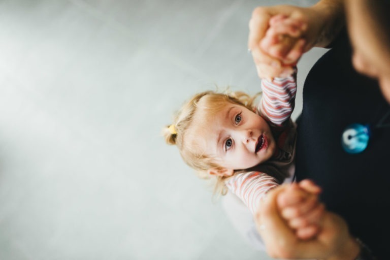 Un jeune enfant aux cheveux blonds coiffés en couettes, vêtu d'une chemise rayée, lève les yeux tout en saisissant les mains d'un adulte. Les mains de l'adulte sont visibles, semblant soulever ou soutenir l'enfant. L'arrière-plan est un sol grisâtre flou. À la Maison du Bonheur, l'enfant a une expression curieuse ou excitée sur son visage.