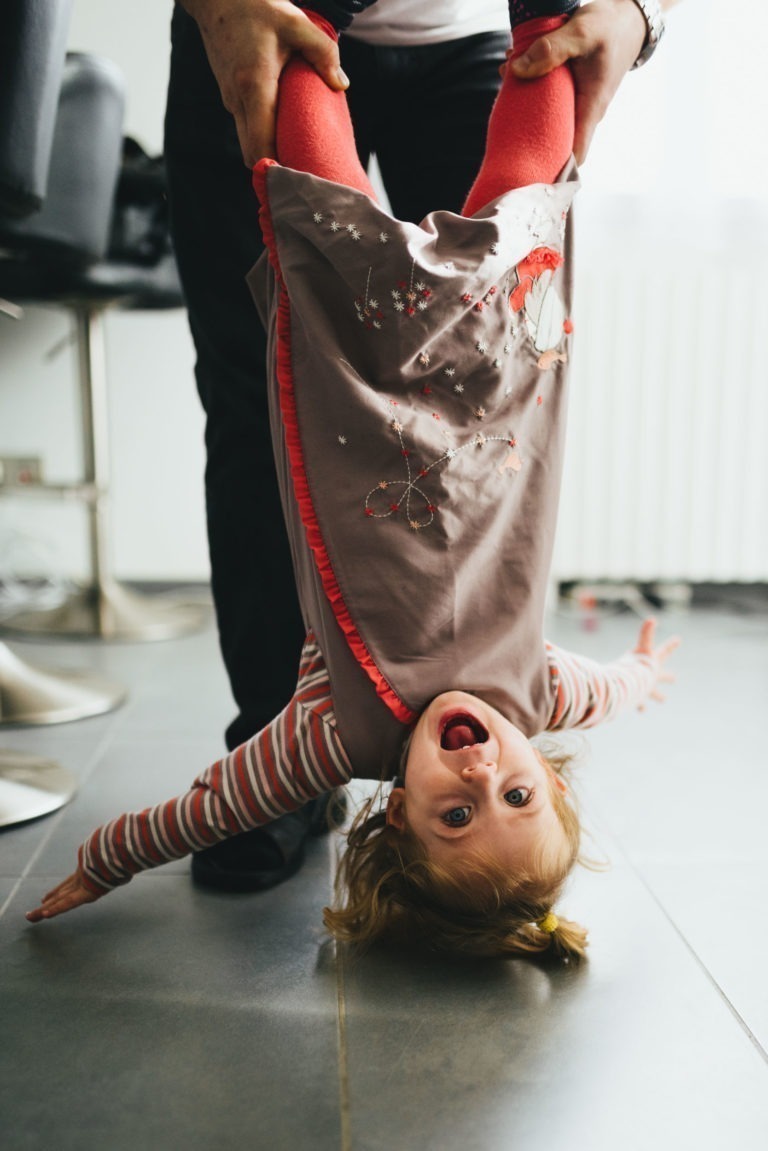 Un jeune enfant aux cheveux châtain clair et à la pince à cheveux est tenu par un adulte, la tête en bas. Vêtu d'une chemise à manches longues rayée, d'une robe marron à motifs rouges et blancs et de collants rouge vif de la Maison du Bonheur, l'enfant joyeux a les yeux écarquillés et un sourire béat. L'arrière-plan comprend une chaise et un sol carrelé.