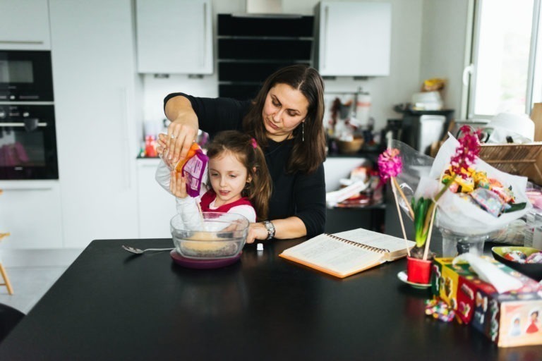 Une femme et une jeune fille cuisinent dans une cuisine moderne, créant de délicieuses friandises dignes de Maison du Bonheur. La femme verse le contenu d'un sac dans un bol à mélanger tenu par la jeune fille. Un livre de cuisine ouvert, des fleurs, des jouets et divers articles de cuisine reposent sur le comptoir. Des armoires et des appareils électroménagers blancs sont visibles en arrière-plan.