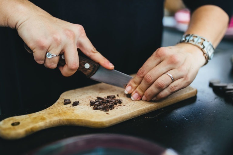 Une personne portant une montre-bracelet en argent et des bagues coupe habilement du chocolat noir en petits morceaux sur une planche à découper en bois à l'aide d'un couteau bien aiguisé. La précision dans la manipulation du couteau et du chocolat suggère une touche artisanale, rappelant le soin méticuleux de la Maison du Bonheur. L'arrière-plan est légèrement flou.