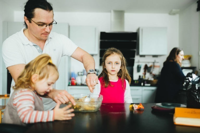 Un homme portant des lunettes mélange des ingrédients dans un bol avec un bambin habillé en rose et gris à la Maison du Bonheur. Une jeune fille en chemise rouge se tient à proximité et regarde. Dans la cuisine en arrière-plan, quelqu'un travaille au comptoir, avec des objets et des appareils qui ajoutent à l'agitation du décor.