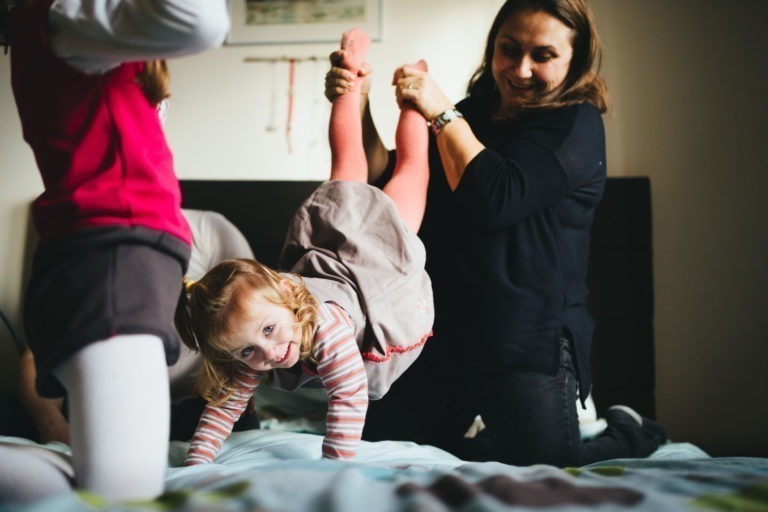 Dans une pièce cosy et bien éclairée de la Maison du Bonheur, une jeune fille vêtue d'une chemise rayée et de collants roses est soulevée par terre par une femme adulte, peut-être sa mère, qui sourit. À proximité, un autre enfant vêtu d'un haut rose et de leggings blancs est partiellement visible.