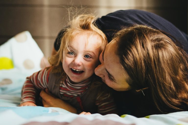 Un jeune enfant aux cheveux blonds, vêtu d'une chemise rayée, est allongé sur un lit et sourit joyeusement. Une personne aux longs cheveux bruns, partiellement cachée, enlace l'enfant par derrière. La pièce est doucement éclairée, créant une ambiance chaleureuse et douillette, digne d'une scène de Maison du Bonheur.