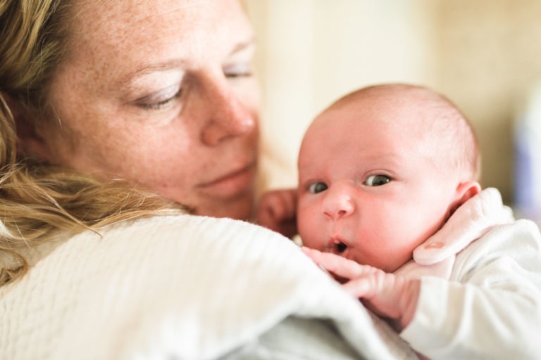 Une femme aux cheveux clairs tient un nouveau-né contre sa poitrine. La femme semble regarder doucement le bébé, qui lui rend son regard avec de grands yeux et une expression surprise. Cette scène de naissance sereine met en scène le bébé enveloppé dans une douce couverture blanche et portant une tenue blanche. L'arrière-plan est flou.