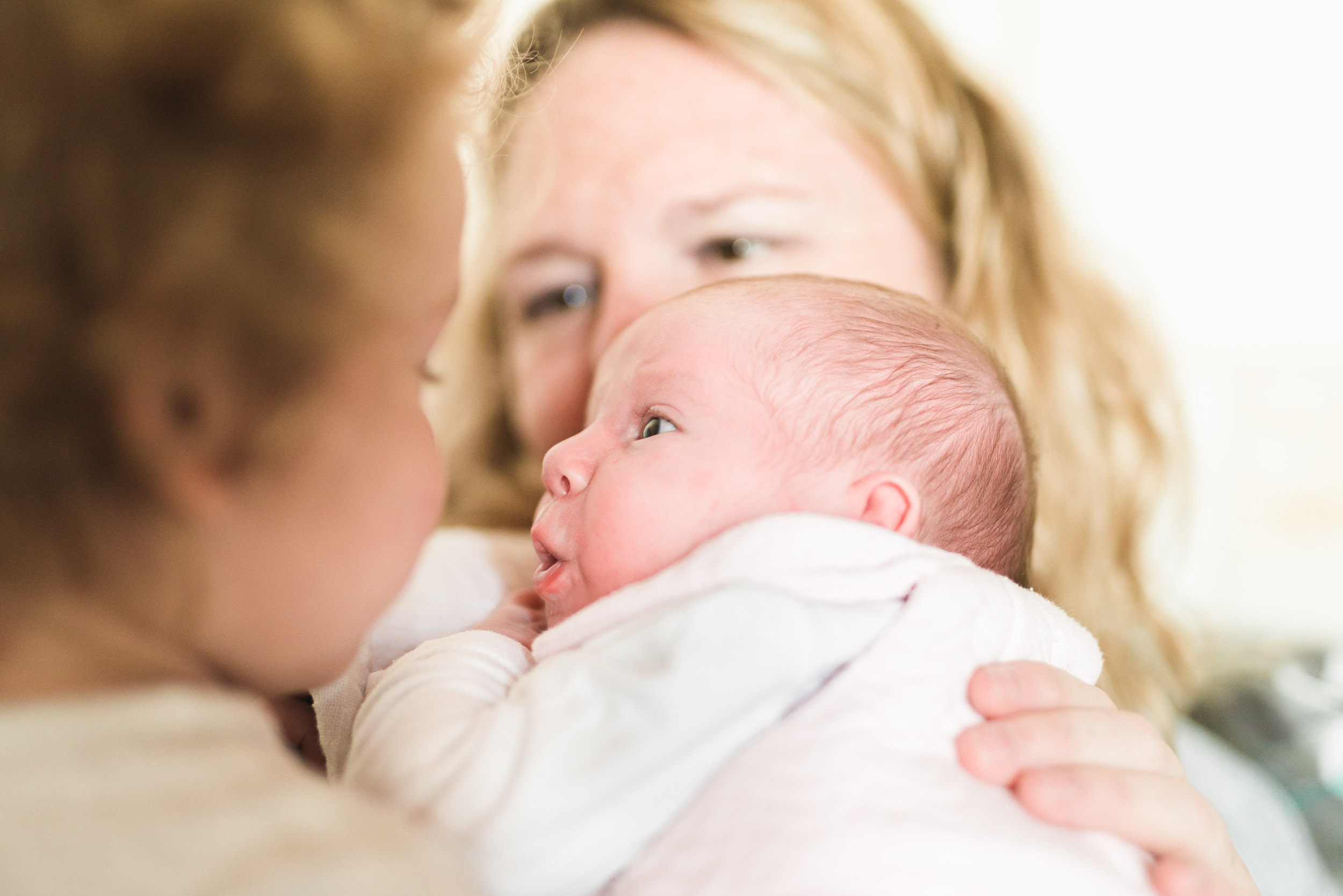 Un bébé enveloppé dans une couverture blanche regarde un enfant qui lui fait face. Derrière le bébé se trouve un adulte aux cheveux blonds et à l'expression douce, qui tient et regarde le nourrisson. La scène de cette naissance sereine apparaît chaleureuse et intime, rayonnant d'affection douce.