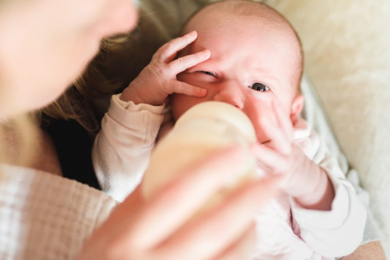 Gros plan d'un nourrisson au visage crispé, nourri au biberon. Le bébé, vêtu d'une combinaison blanche, porte une main à son visage tout en buvant au biberon tenu par un adulte. L'arrière-plan est légèrement flouté et la scène semble tendre et intime, capturant un moment de naissance sereine.