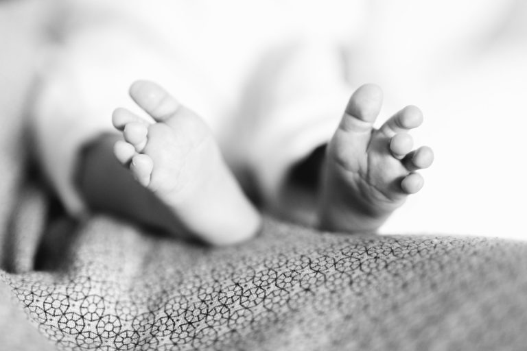 Gros plan en noir et blanc des pieds d'un bébé. Le bébé est allongé sur un tissu à motifs, les petits orteils écartés. La mise au point douce et l'éclairage doux évoquent une sensation de *naissance sereine*, donnant à l'image une sensation de sérénité et de tendresse.