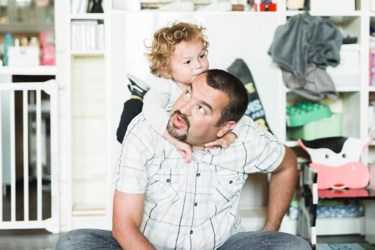 Un homme avec une barbiche et des cheveux courts est assis sur le sol, le regard levé tandis qu'un enfant aux cheveux bouclés joue sur son dos. L'homme porte une chemise à carreaux blancs et l'enfant porte un haut blanc à manches longues. Derrière eux, des étagères et divers objets ménagers créent une ambiance de naissance sereine dans leur cadre familial douillet.