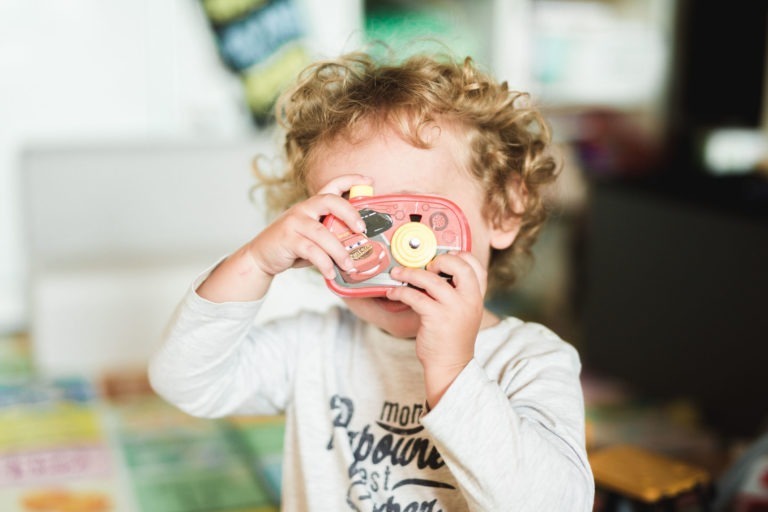 Un bambin aux cheveux blonds bouclés tient un appareil photo jouet devant son visage comme s'il prenait une photo. L'enfant porte un t-shirt blanc à manches longues avec le texte imprimé « naissance sereine ». L'arrière-plan est flou, montrant des tapis de jeu et des étagères colorés, suggérant une aire de jeux intérieure.