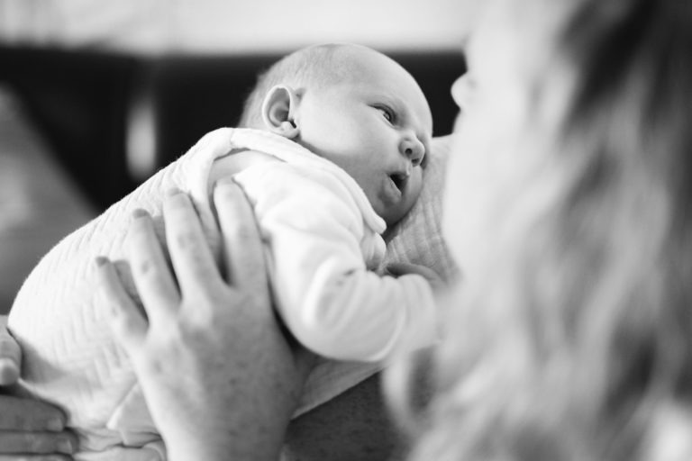 Image en noir et blanc d'un bébé appuyé contre la poitrine d'un adulte. Le bébé, vêtu d'une tenue claire, regarde vers le haut avec de grands yeux. L'adulte, vu de profil avec les cheveux flous au premier plan, tient doucement le bébé, évoquant un sentiment de réconfort et de lien, un véritable moment de naissance sereine.